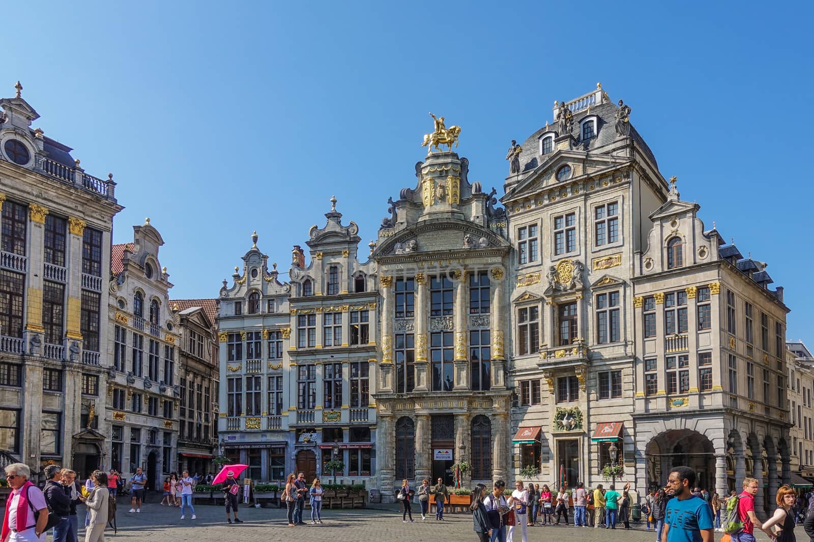 Brussels, Belgium - June 22, 2019: Grand Place with tourists and beige stone facades, gables and golden statues of southwest side against blue sky. Most famous, Arbre d’or, Etoile, Le Cygne,