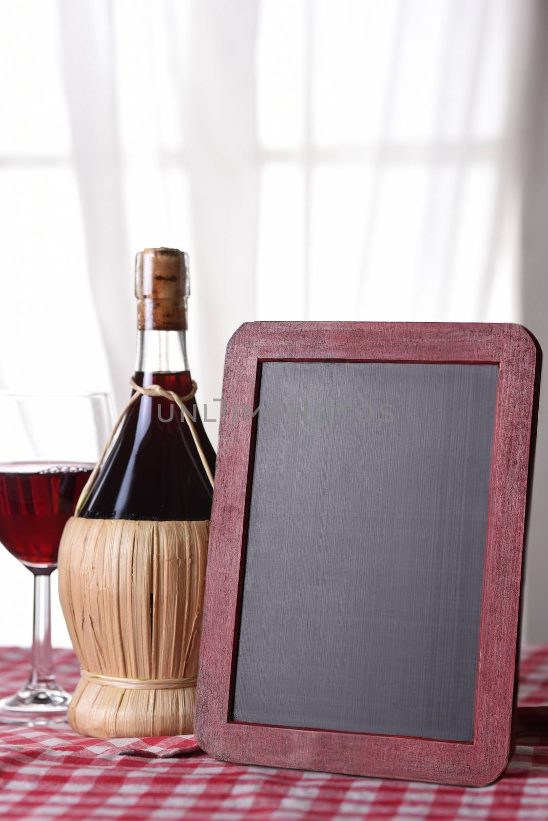 A basket bottle of Chianti wine on a red checkered tablecloth with a blank wine menu board. 
