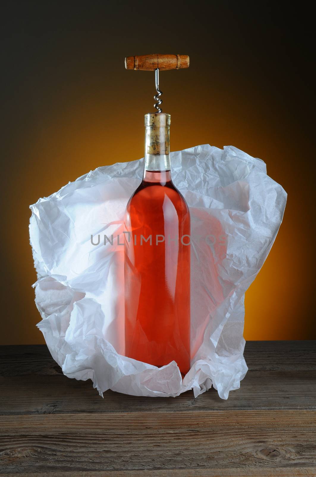 A bottle of blush wine wrapped in tissue paper, on a rustic wood surface and a light to dark warm background.
A vintage cork screw is inserted in the bottle.