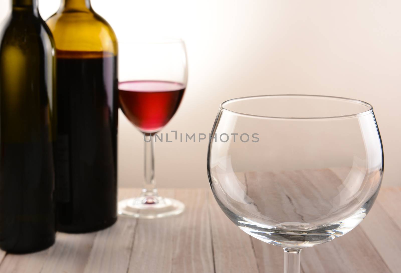 Closeup of an empty wineglass with bottles and full glass in the background. Horizontal still life with warm tones and light to dark background lighting.