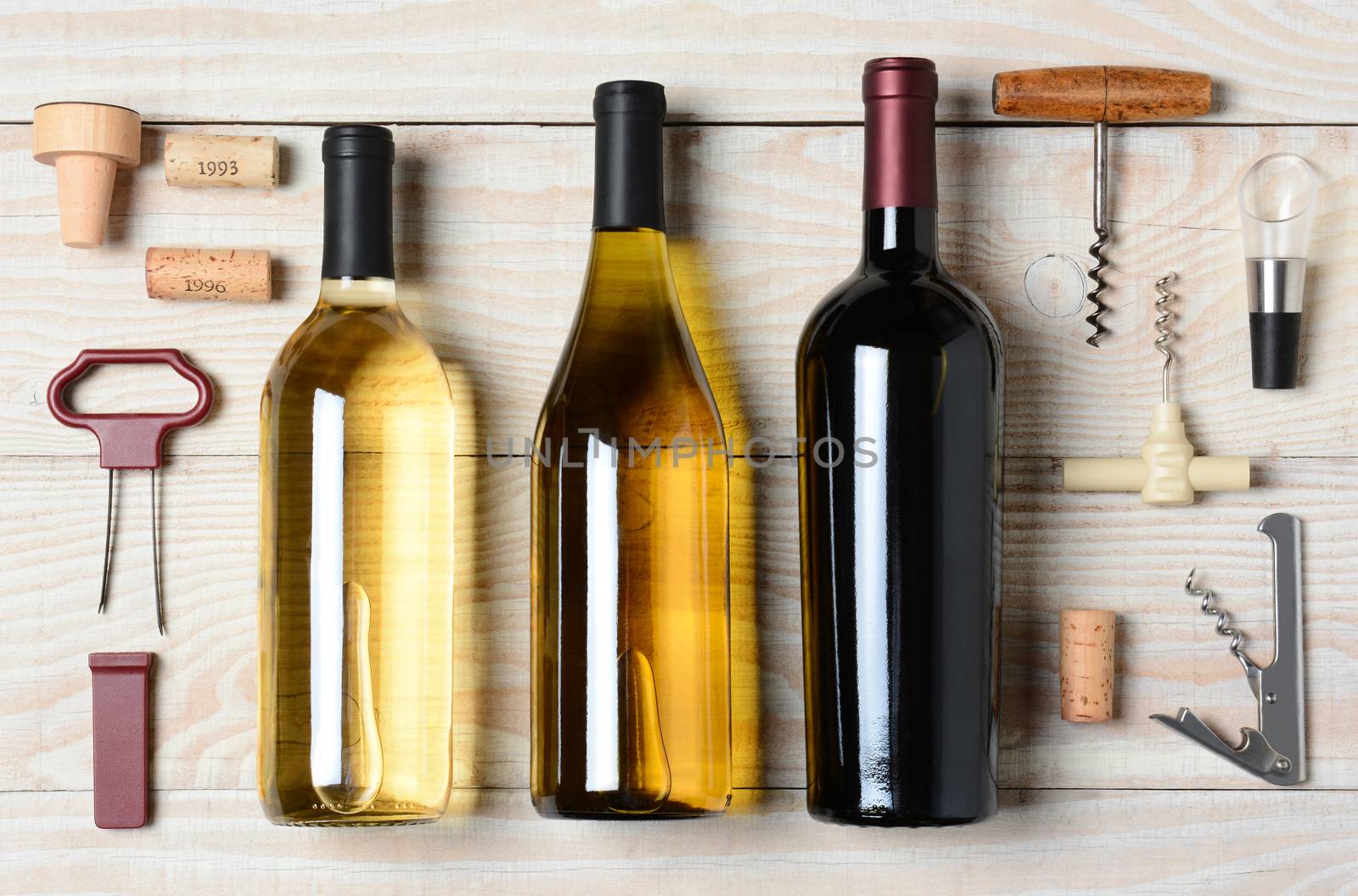High angle shot of three wine bottles surrounded by accessories such as corkscrews, stoppers, pourers and corks. Horizontal format on a rustic white wood table.