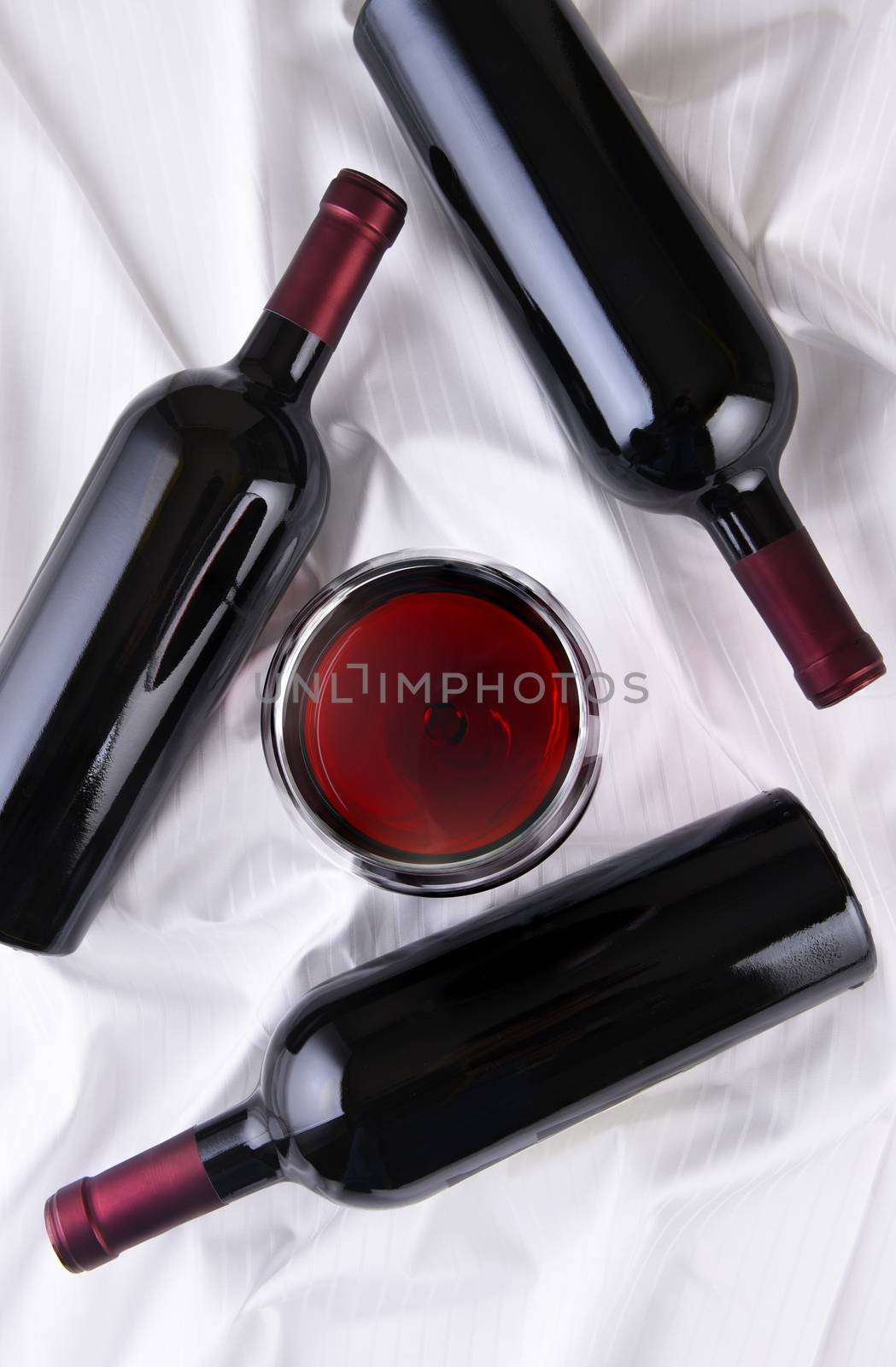Wine glass in the center of three bottles laying on their sides on white fabric surface. Vertical orientation.