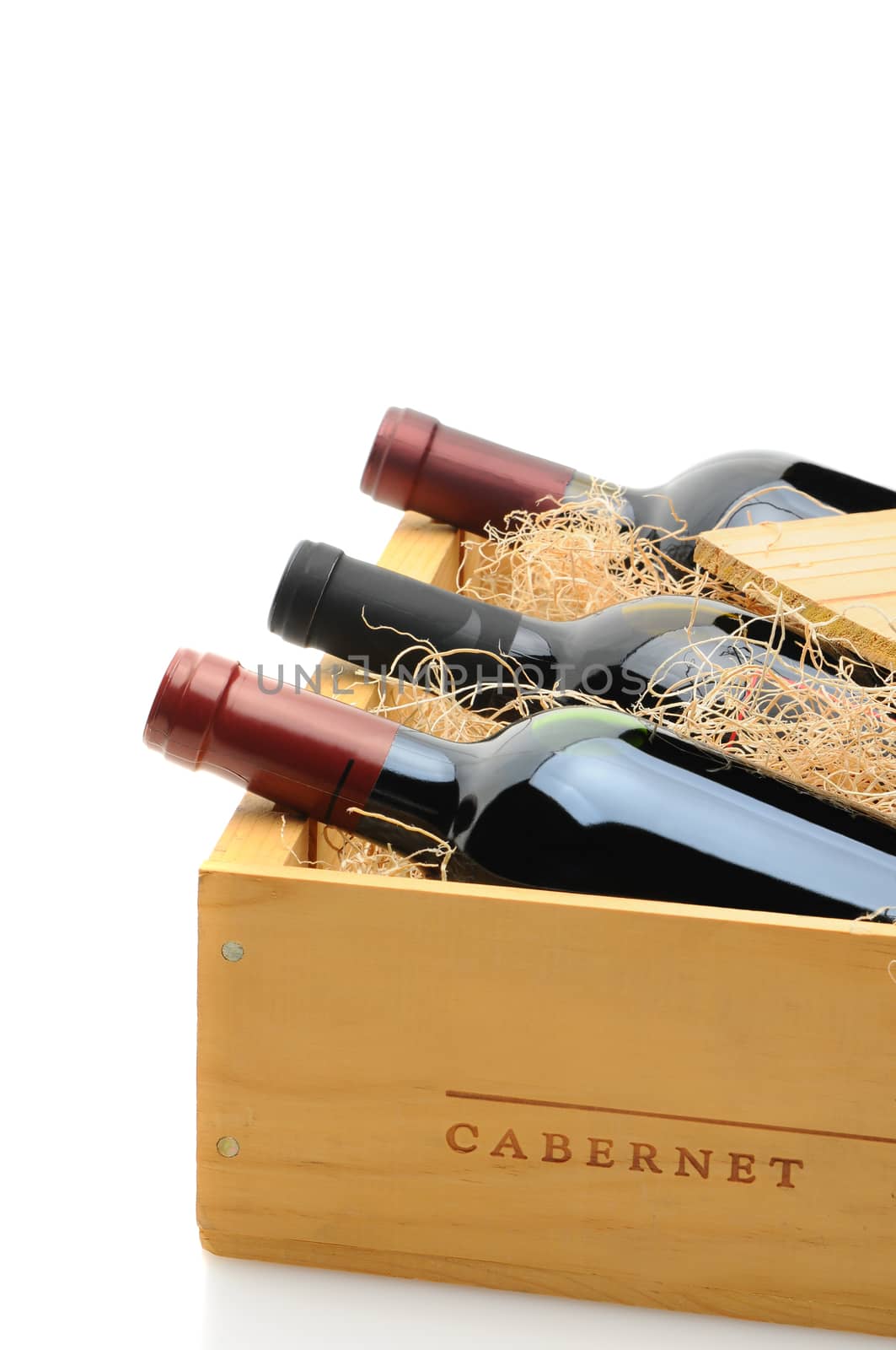 Closeup of three red wine bottles in a wooden shipping crate. The crate is partially open with excelsior packing. Vertical format over a white background with reflection.