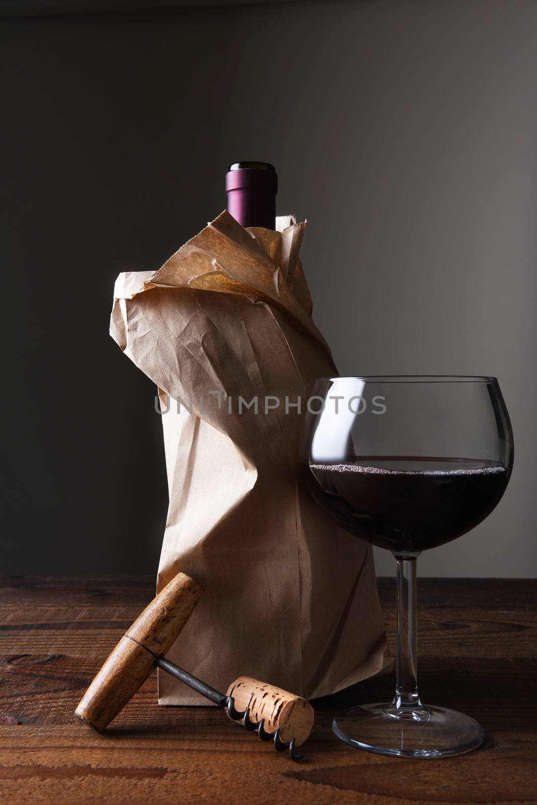 Closeup of a wine bottle in a paper bag on a wooden table with a glass and corkscrew.
