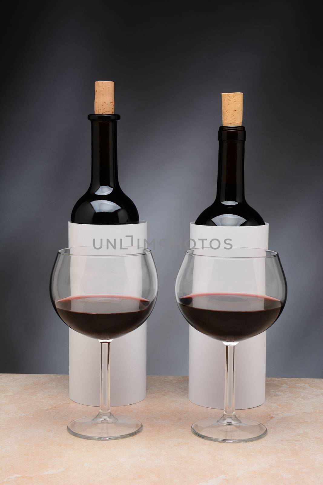 Two different wine bottles and wine glasses set up for a blind wine tasting. The bottles are covered by blank cylinders to hide the label. Vertical format. Wine glasses are partially filled with red wine.