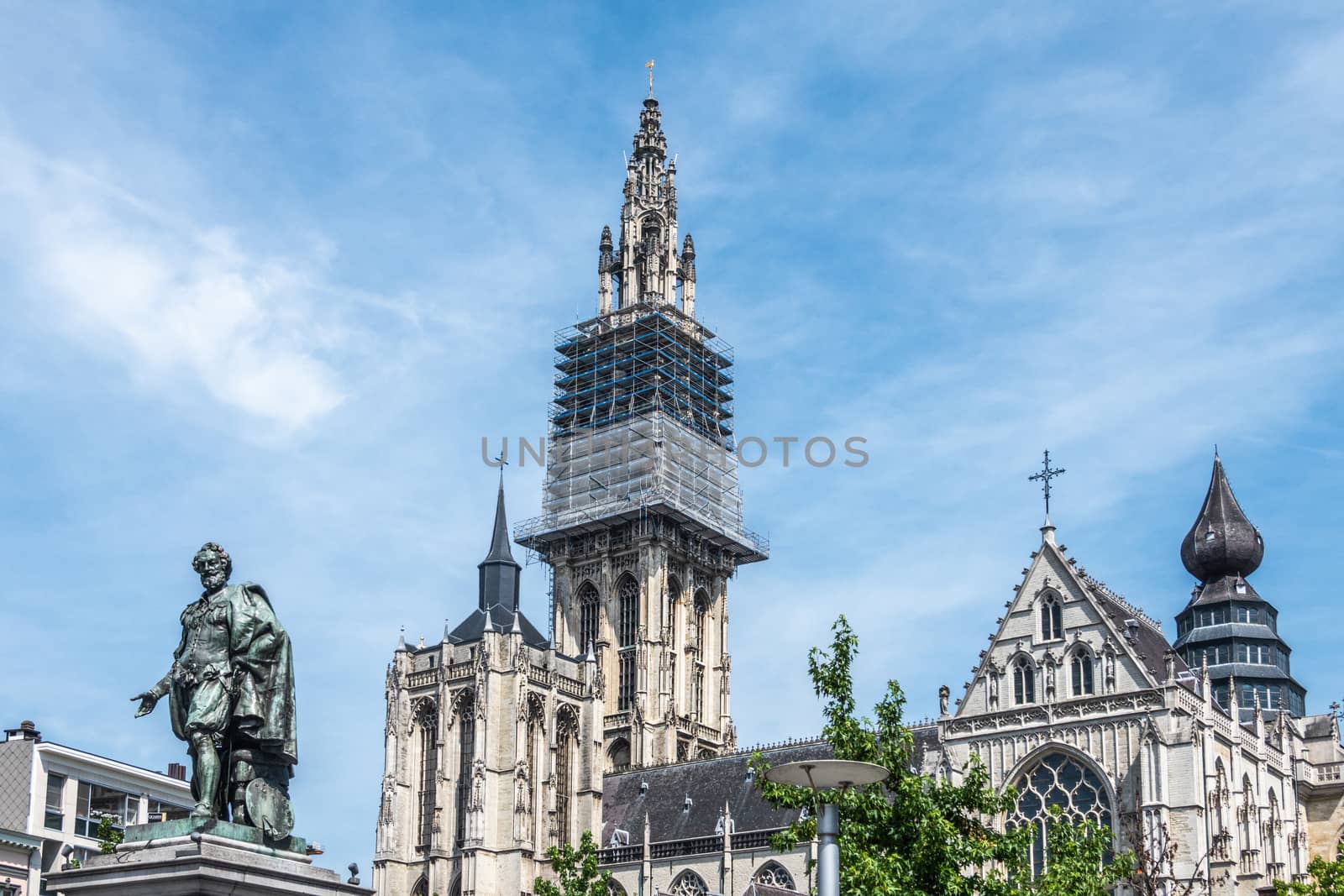 Antwerpen, Belgium - June 23, 2019: Bronze Peter Paul Rubens statue fronts towers and nave of Notre Dame, Onze-Lieve-Vrouw, cathedral against light blue sky. Some green foliage.