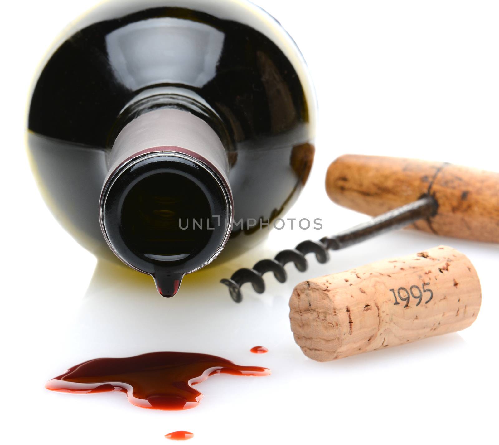 Closeup of a red wine bottle with a drip and wine spill in the foreground. A cork screw and cork to one side on a white background with slight reflection. Shallow depth of field with focus on the drip.