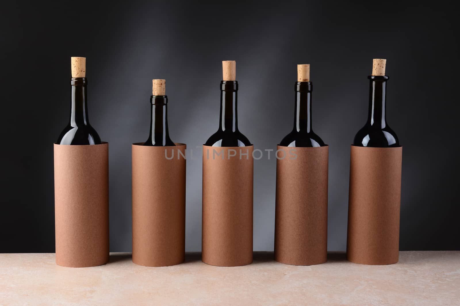 Five different wine bottles set up for a blind wine tasting. The bottles have the corks partially removed and are covered by blank cylinders to hide the label. Horizontal format.