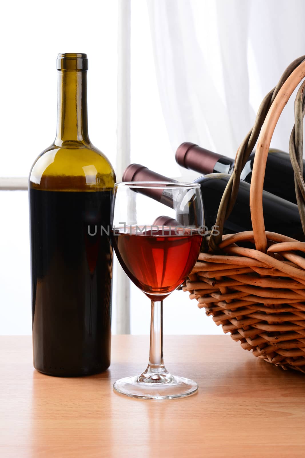 Wine still life with a basket of bottles and a glass of red wine in front of a window with curtains. Vertical format.