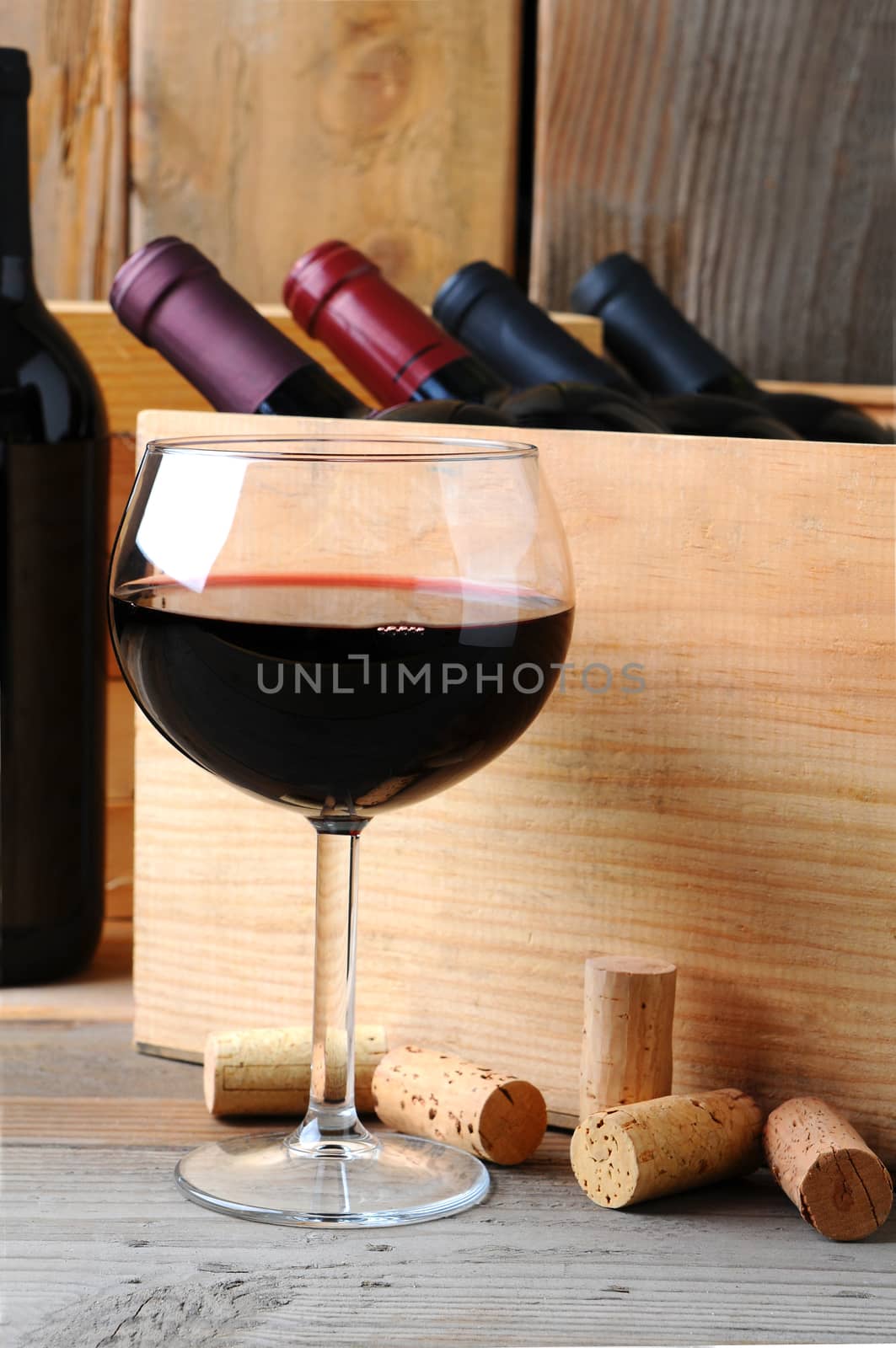 A glass of red wine in front of a wooden case of wine bottles on a rustic wood background. Vertical Format.