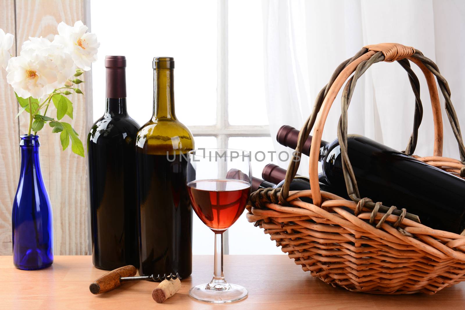 Wine still life with a basket of bottles and a glass of red wine and white roses in a vase on a table in front of a window with curtains. Horizontal format.