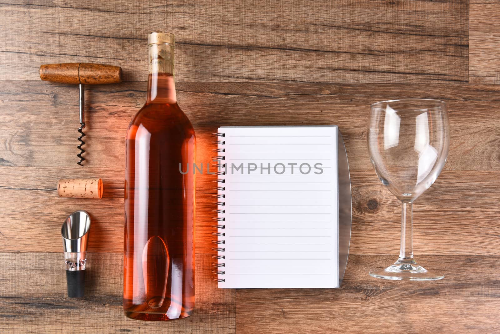 A high angle view of a wine bottle tasting notes notebook, cork screw, and wine glass on a wood table.