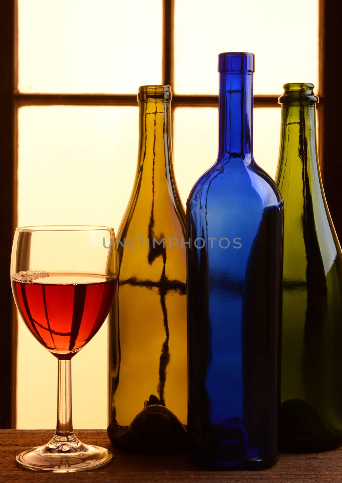 A wine still life with warm tones. Three different wine bottles and a glass of red wine in front of a window with warm sunlight.