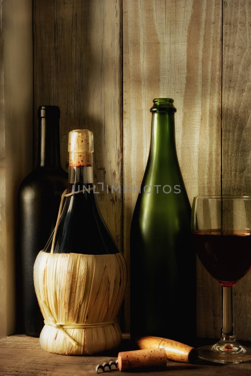 A wine still life with warm light from a window on the side. Three bottles, a wine glass and cork screw in a rustic setting, Vertical format, 