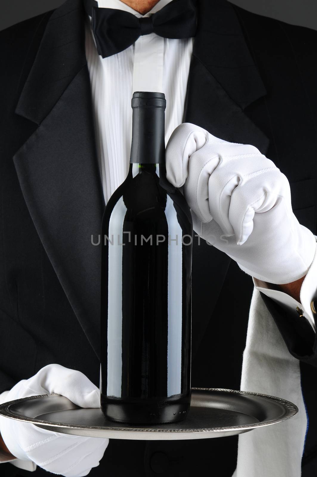 Closeup of a waiter wearing a tuxedo and white gloves holding a wine bottle on a serving tray. Vertical format. The man is unrecognizable.