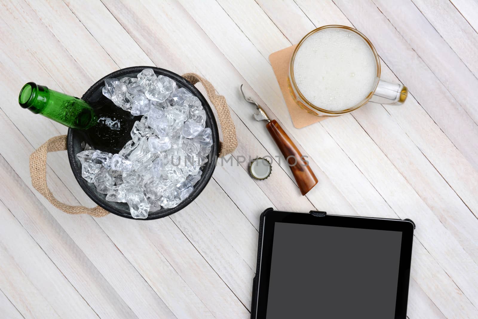 Overhead shot of an ice bucket with an opened beer bottle, a mug of beer and tablet computer for electronic ordering on a rustic white wood table. Horizontal format with copy space.