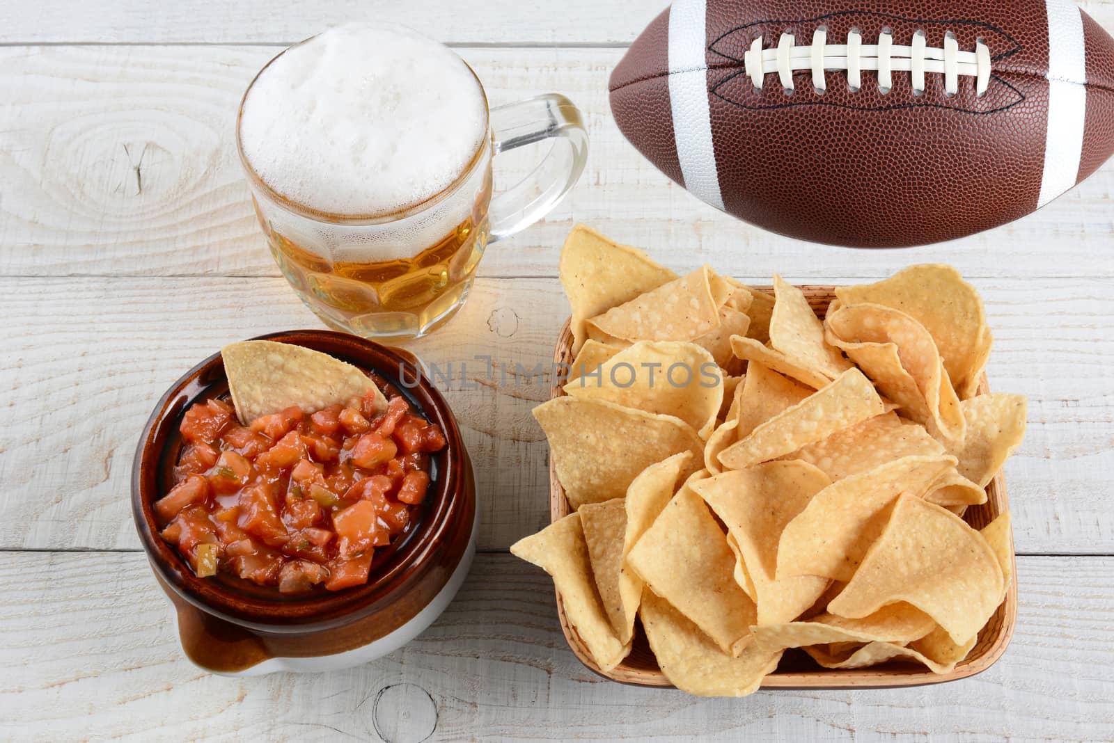 High angle shot of a bowl of corn chips a crock full of fresh salsa a mug of beer and an American football on a whitewashed rustic wood table. Horizontal format. 