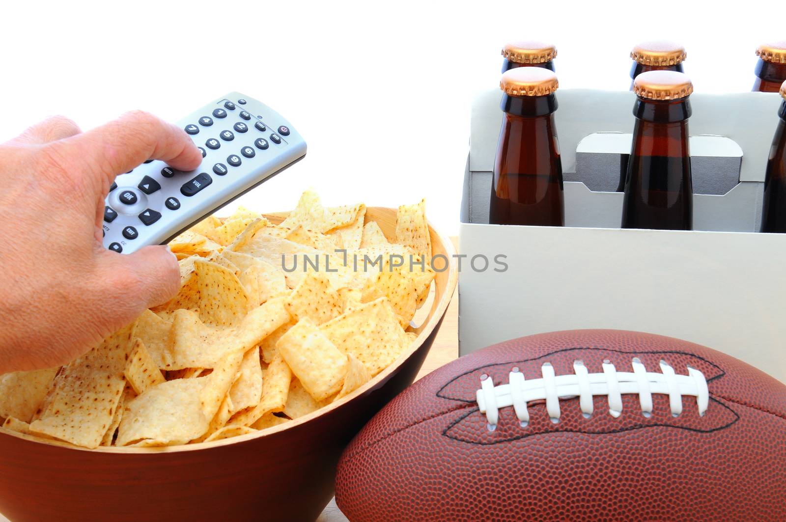 Closeup of a man's hand holding a TV remote with a bowl of chips six pack of beer and football isolated on white. Horizontal Format.