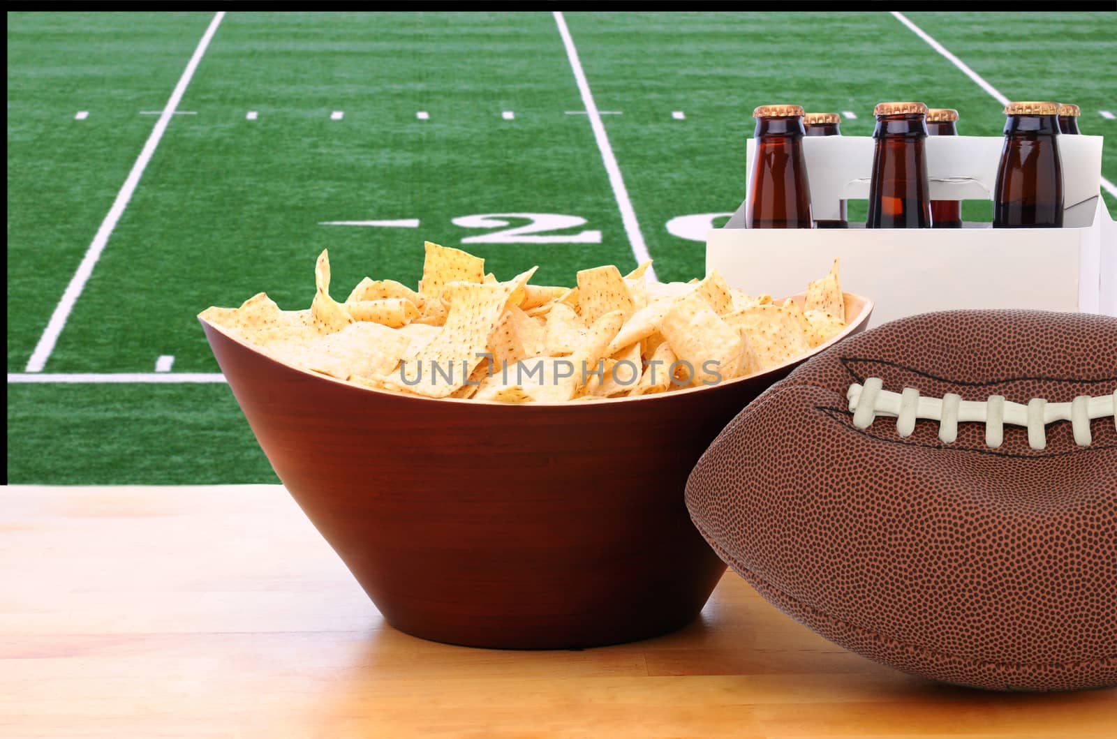 A deflated football and Six Pack of Beer and bowl of chips on a table in front of a big screen TV with a Football field. Great for Super Bowl themed projects. Horizontal format.