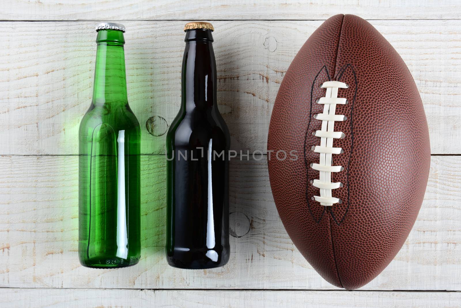 Two beer bottles and an American style football on a rustic whitewashed wood surface. Horizontal format. One green bottle and one brown, both without labels.