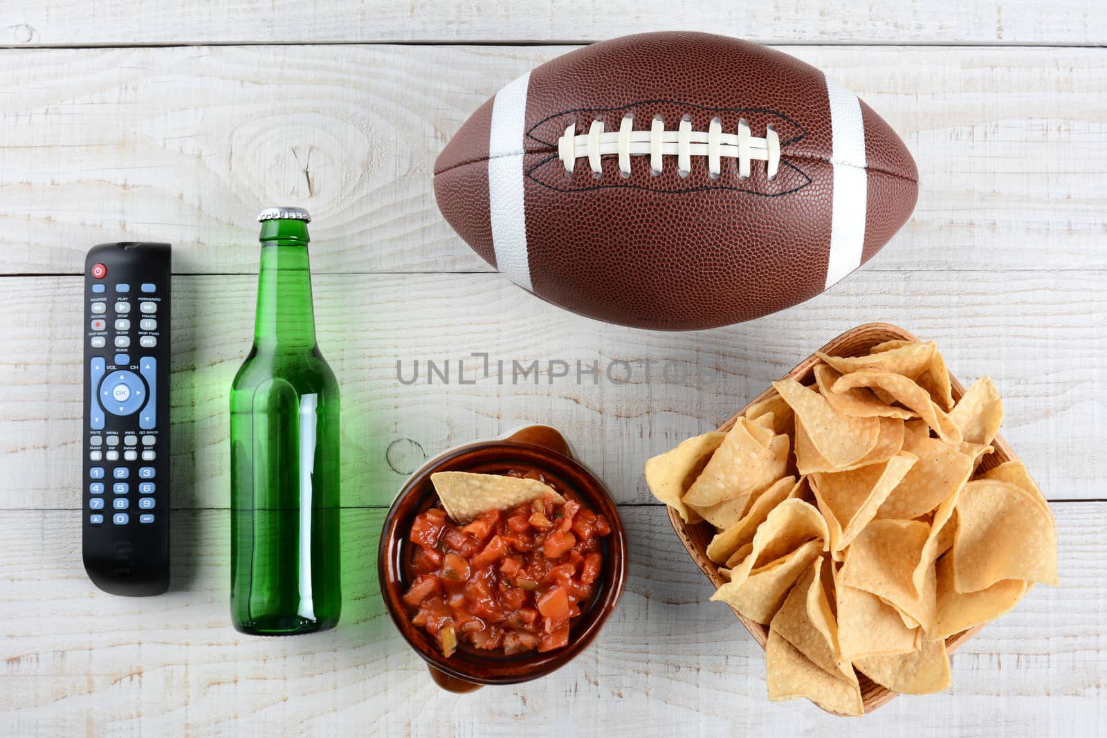 TV Remote, beer bottle, bowl of chips with salsa and an American style football on a rustic whitewashed wood surface. Horizontal format. Great for Super Bowl party themed projects.