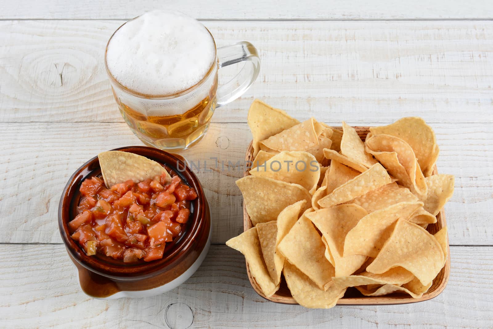 High angle shot of a bowl of corn chips a crock full of fresh salsa and mug of beer on a whitewashed rustic wood table. Horizontal format. 