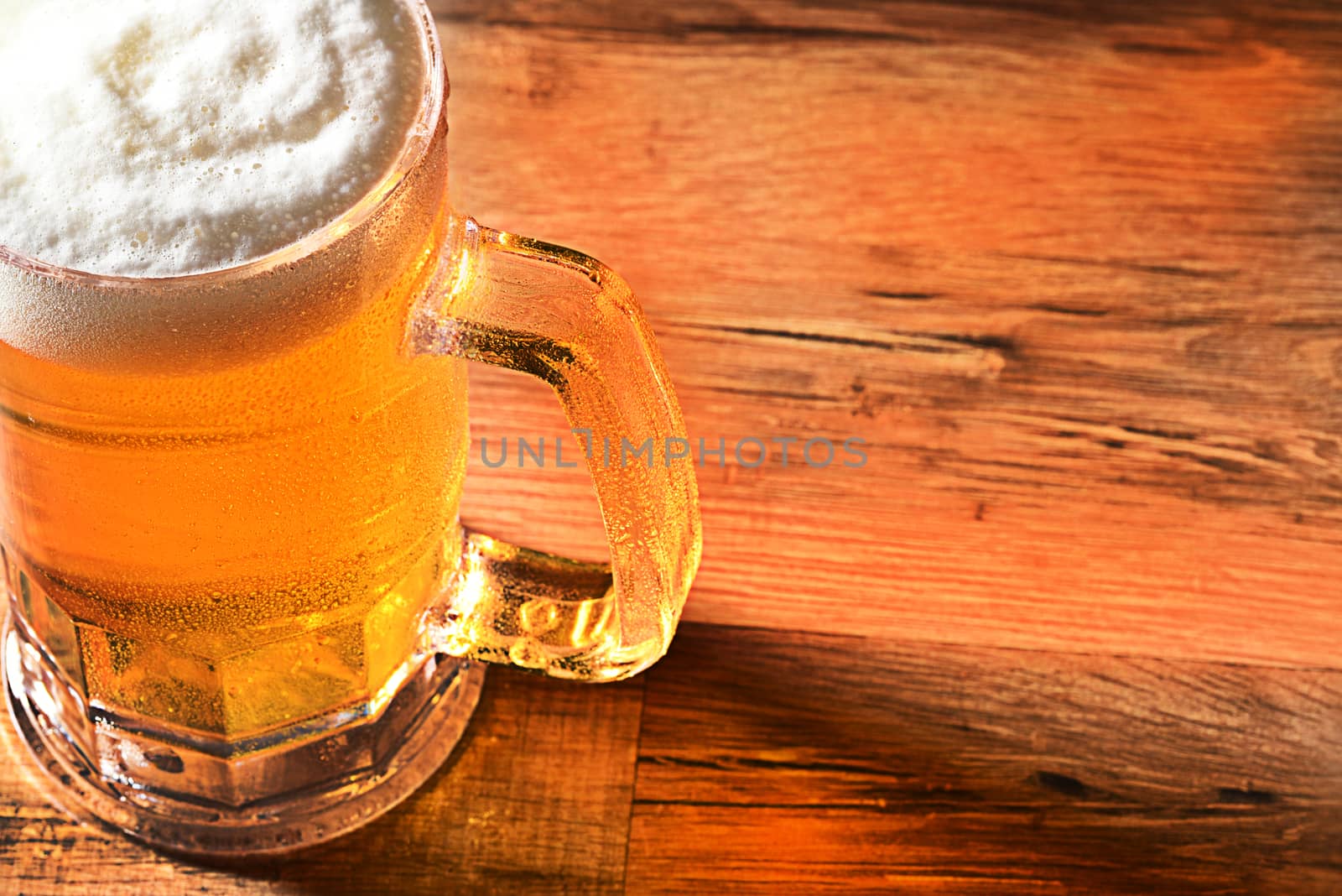 Closeup of a cold beer mug with a foamy head and copy space. High angle view with warm side light and reflections on wood table.