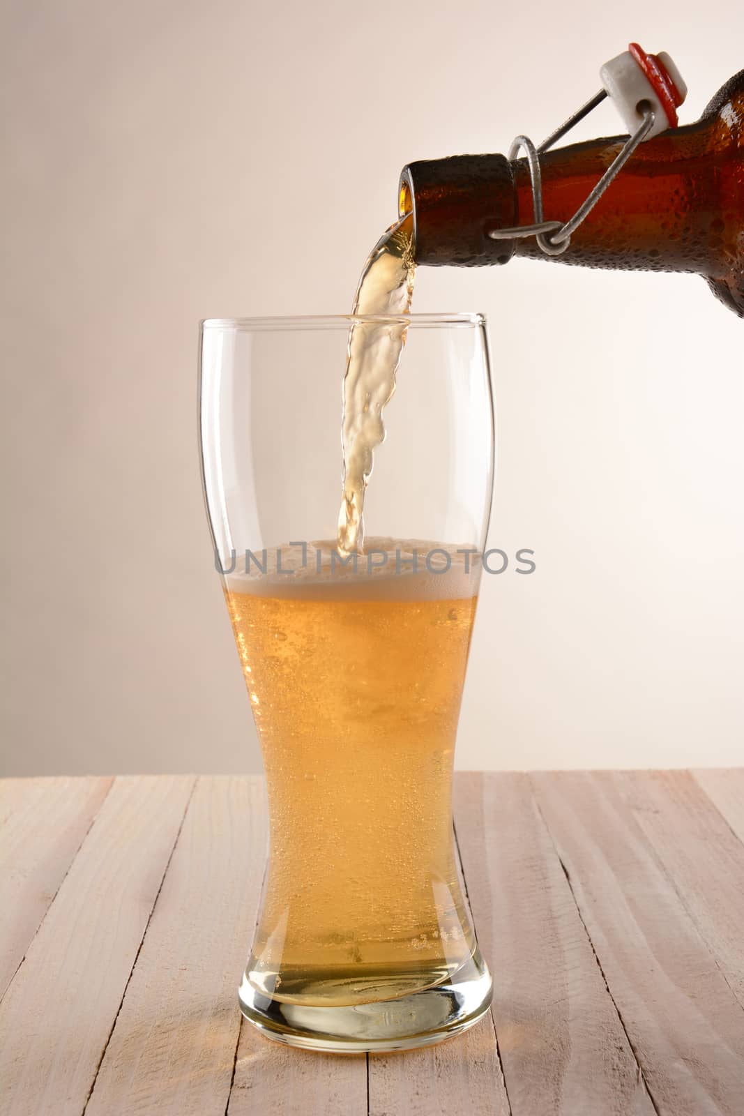 A brown swing top bottle of beer pouring into a glass. The partially filled glass is on a wood table with a light to dark background.