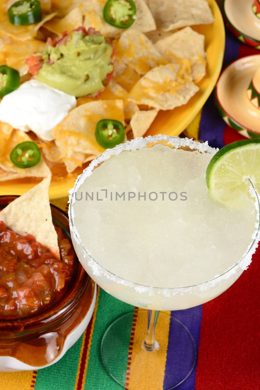 High angle view of a margarita cocktail surrounded by nachos, chips, salsa on a bright Mexican, table cloth. Vertical format. Perfect for Cinco de Mayo projects.