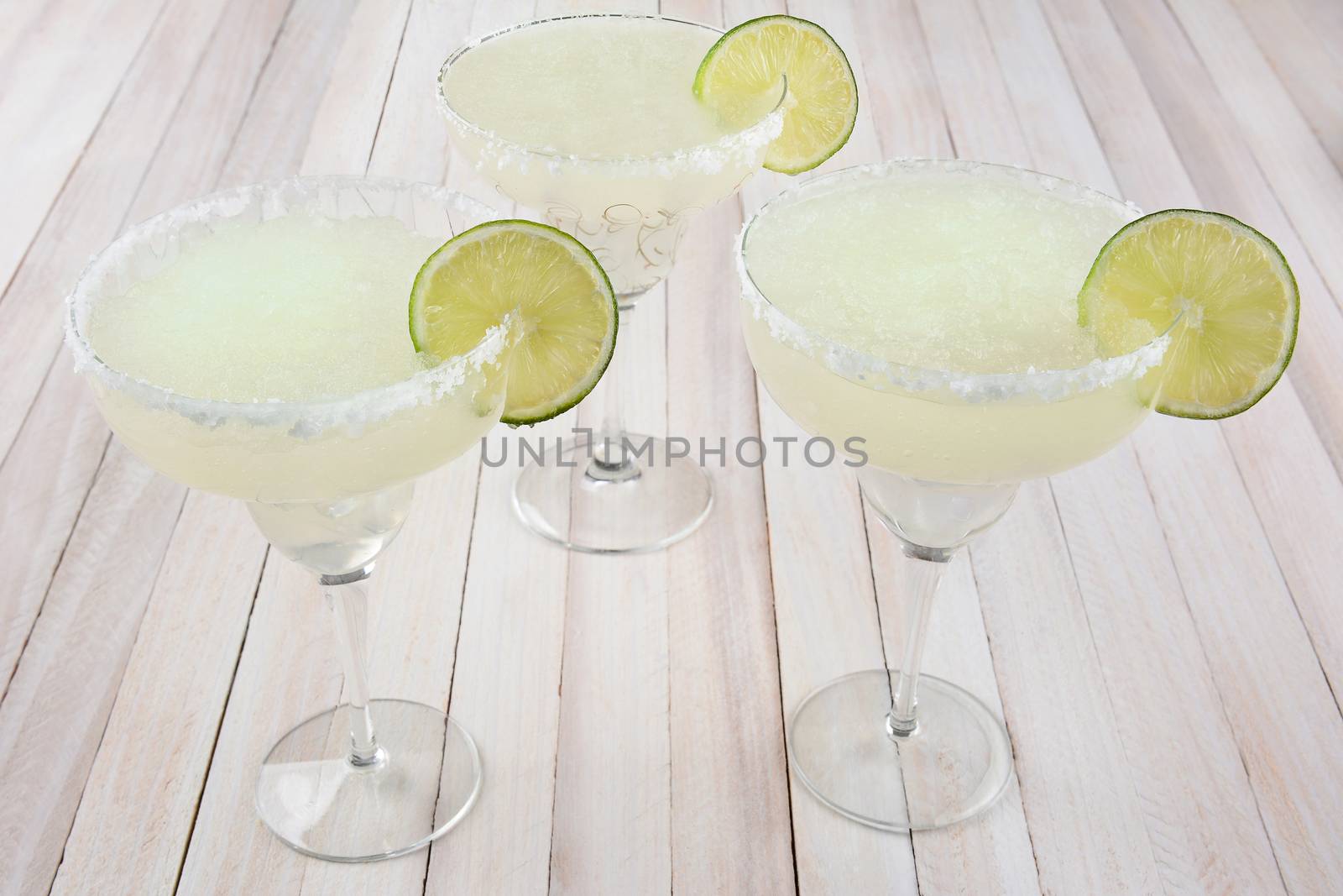 High angle view of three margarita cocktails on a rustic white wood table. Horizontal format.