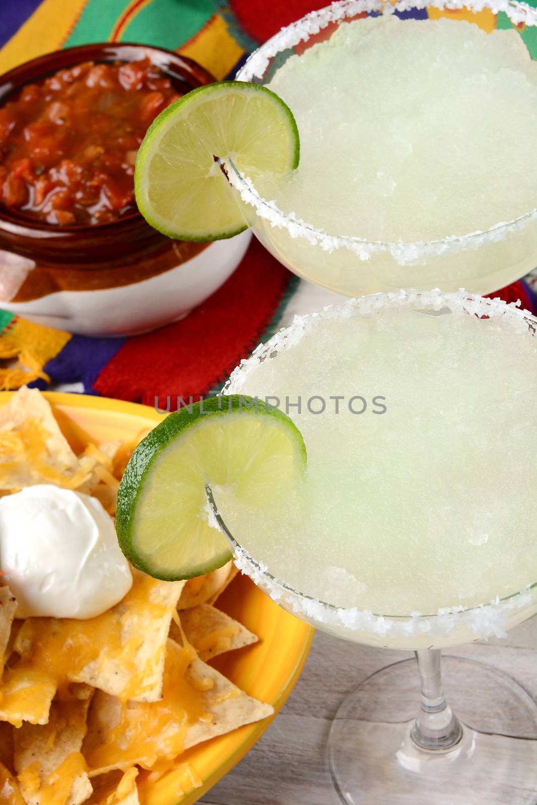 High angle view of two margarita cocktails for a Cinco de Mayo celebration. Surrounded by nacho chips and salsa on a bright Mexican table cloth.