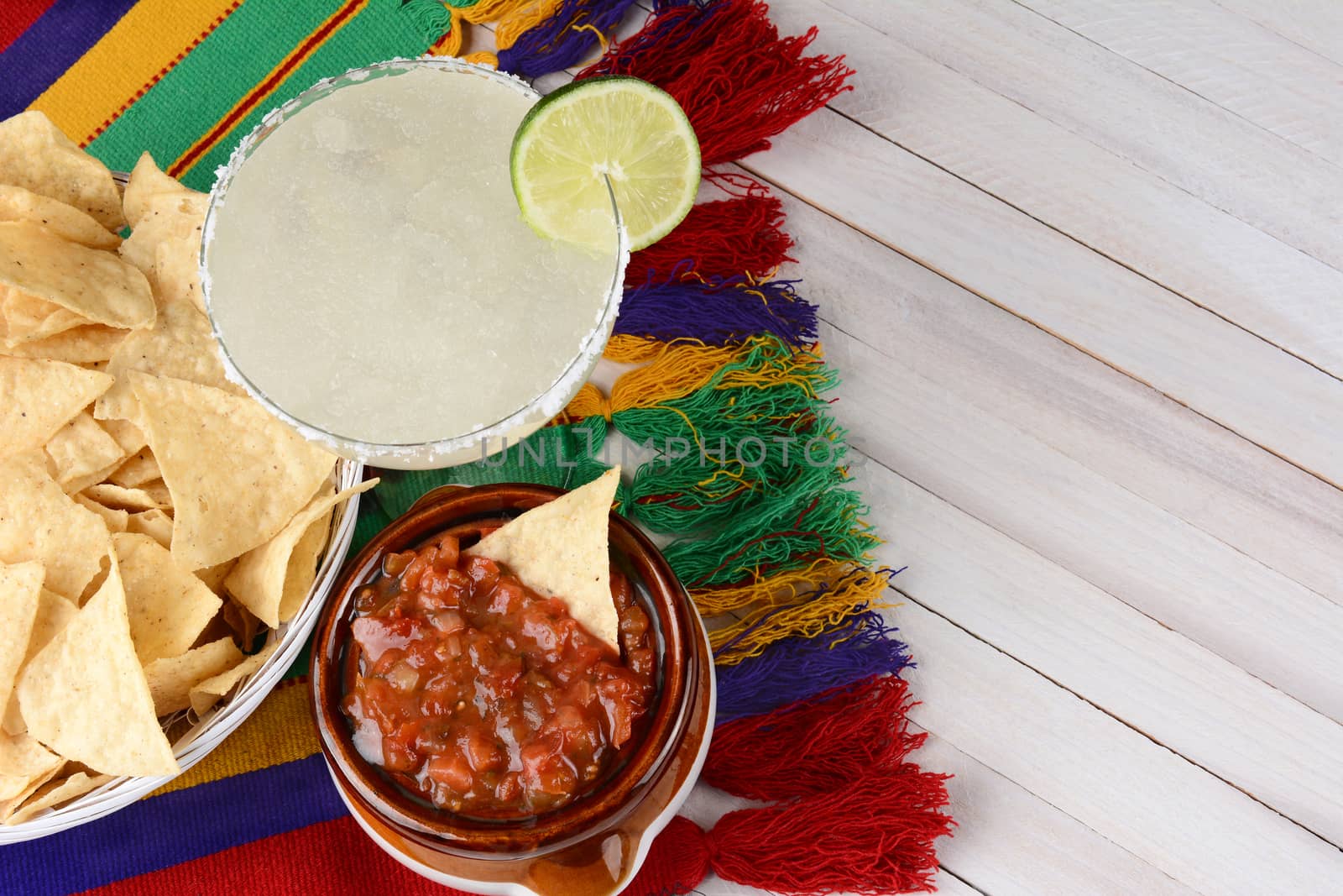 High angle view of a margarita cocktail with chips and salsa on a white rustic wood table. Horizontal format with copy space.