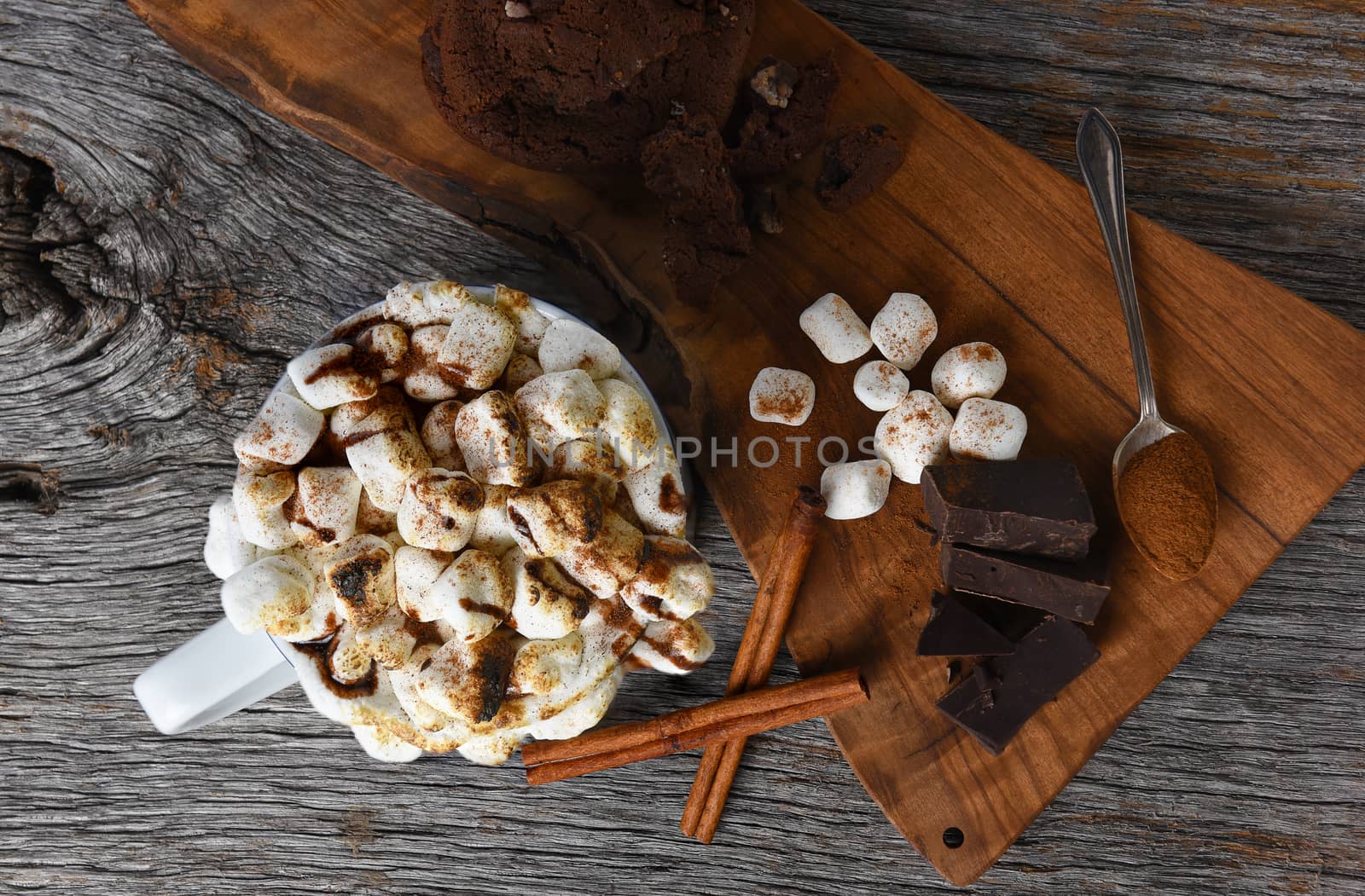 Overhead shot of a mug of hot cocoa with toasted marshmallows next to a cutting board with chocolate chunks cinnamon sticks and cookies. 