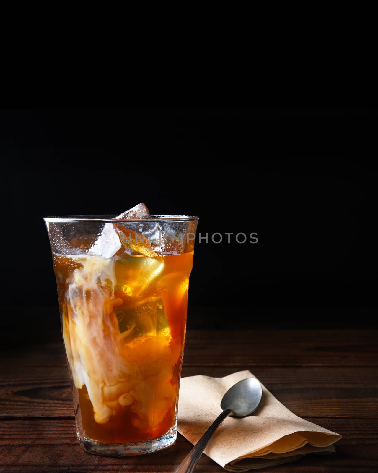 Closeup of a glass of iced coffee on a dark wood table. Fresh poured cream is permeating through the glass with a napkin and spoon lay next to the glass. Vertical with copy space.