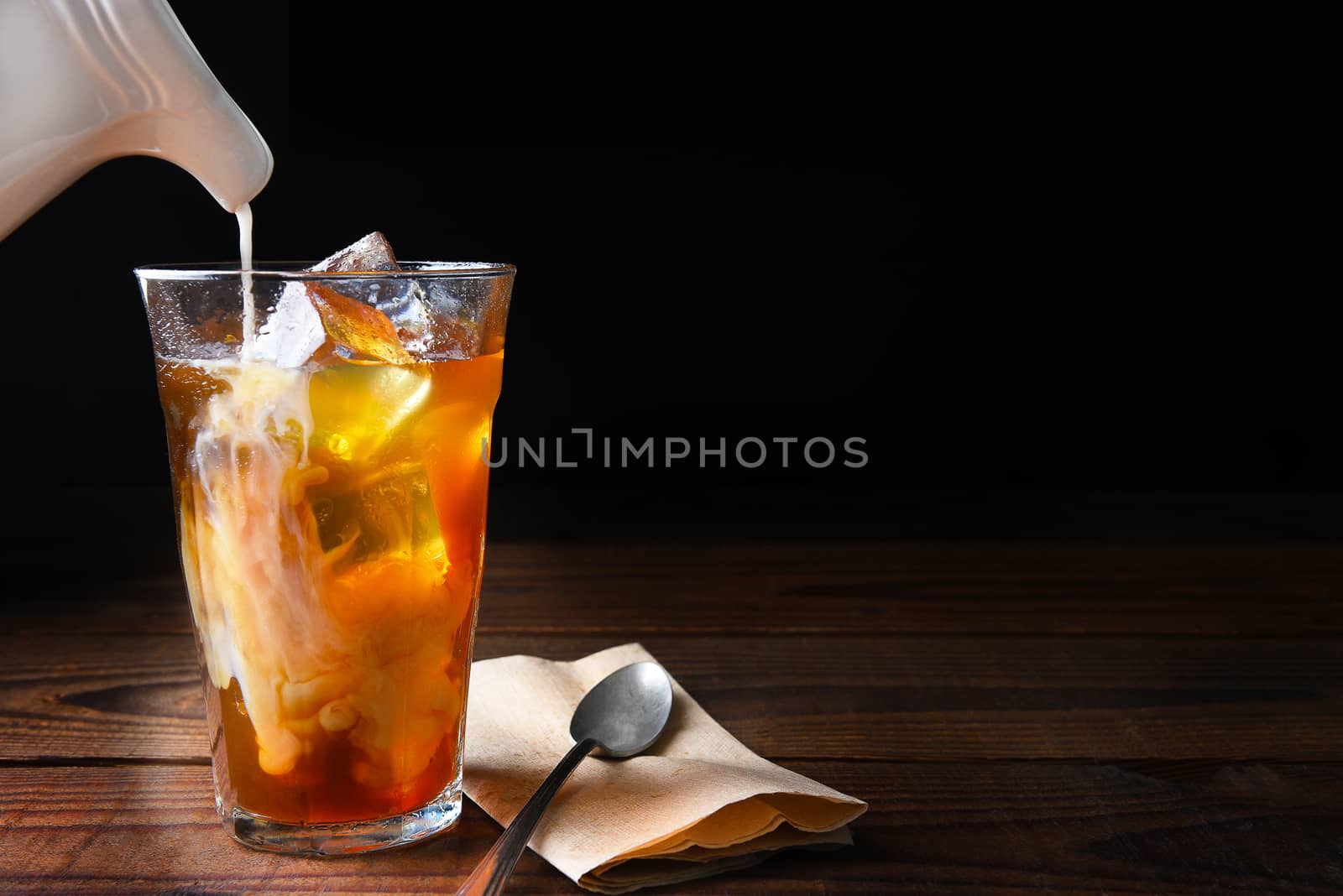 Closeup of a glass of iced coffee on a dark wood table. Fresh pouring cream is permeating through the glass with a spoon and napkin next to the glass. Horizontal on a black background with copy space.