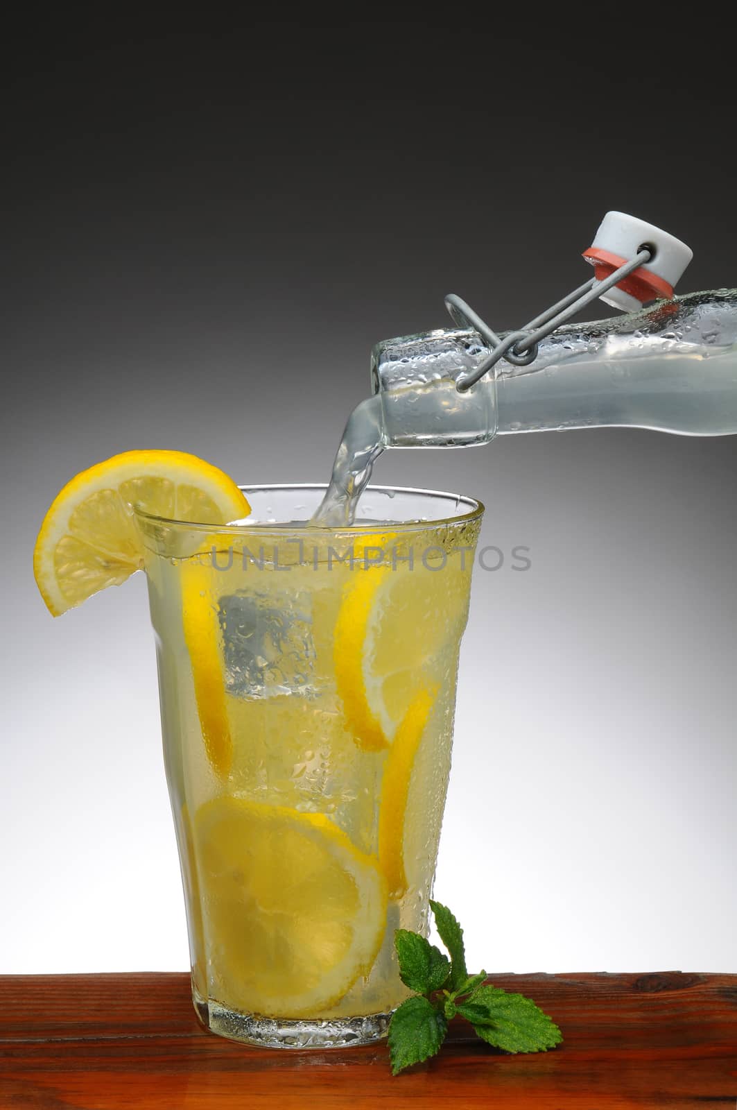 Lemonade pouring into a glass from a bottle. Glass is on a rustic wooden table with a light to dark gray background.