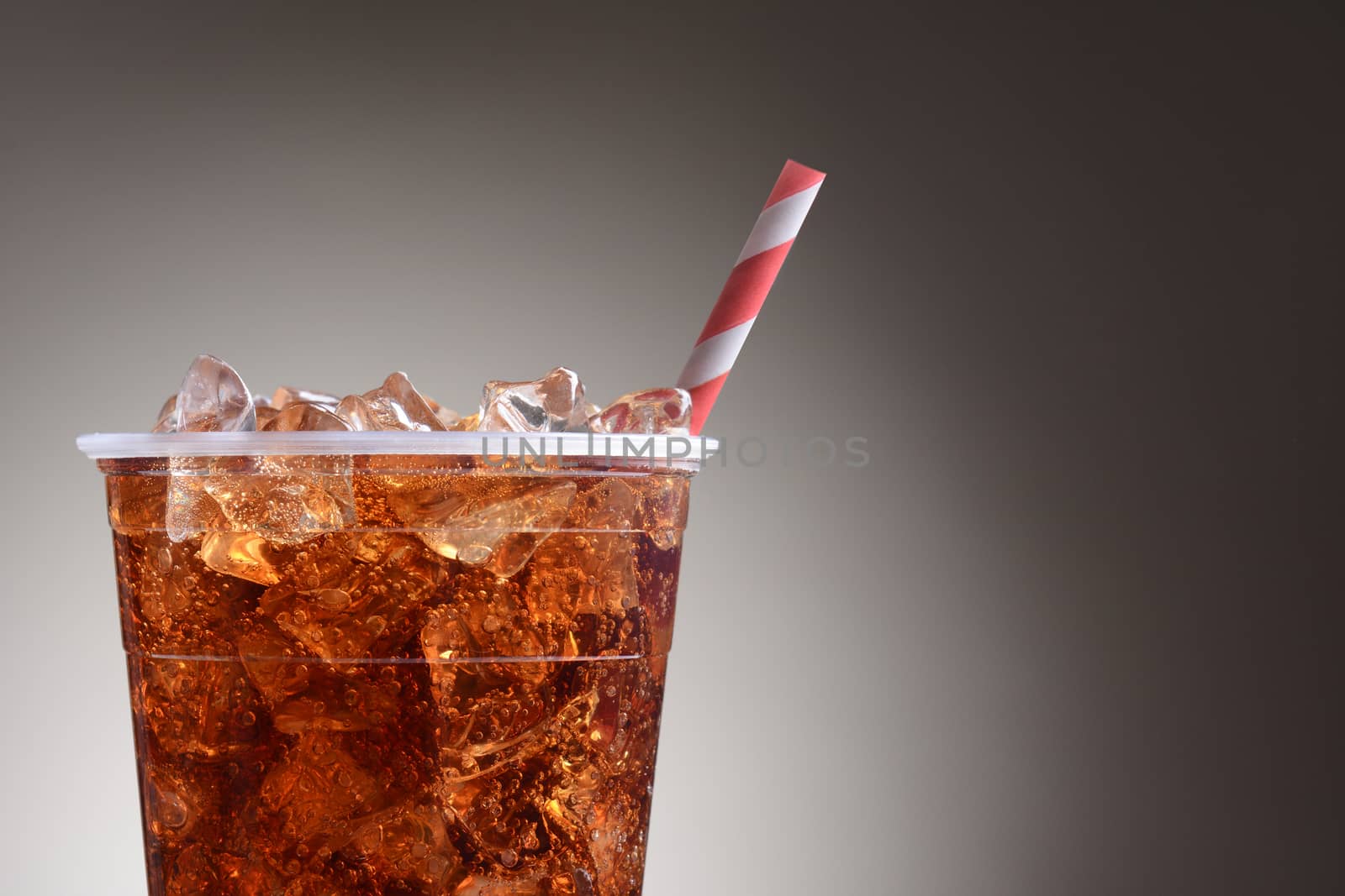 A clear plastic cup filled with ice and cola. Only half the cup is shown with a red and white striped straw over a light to dark gray background.