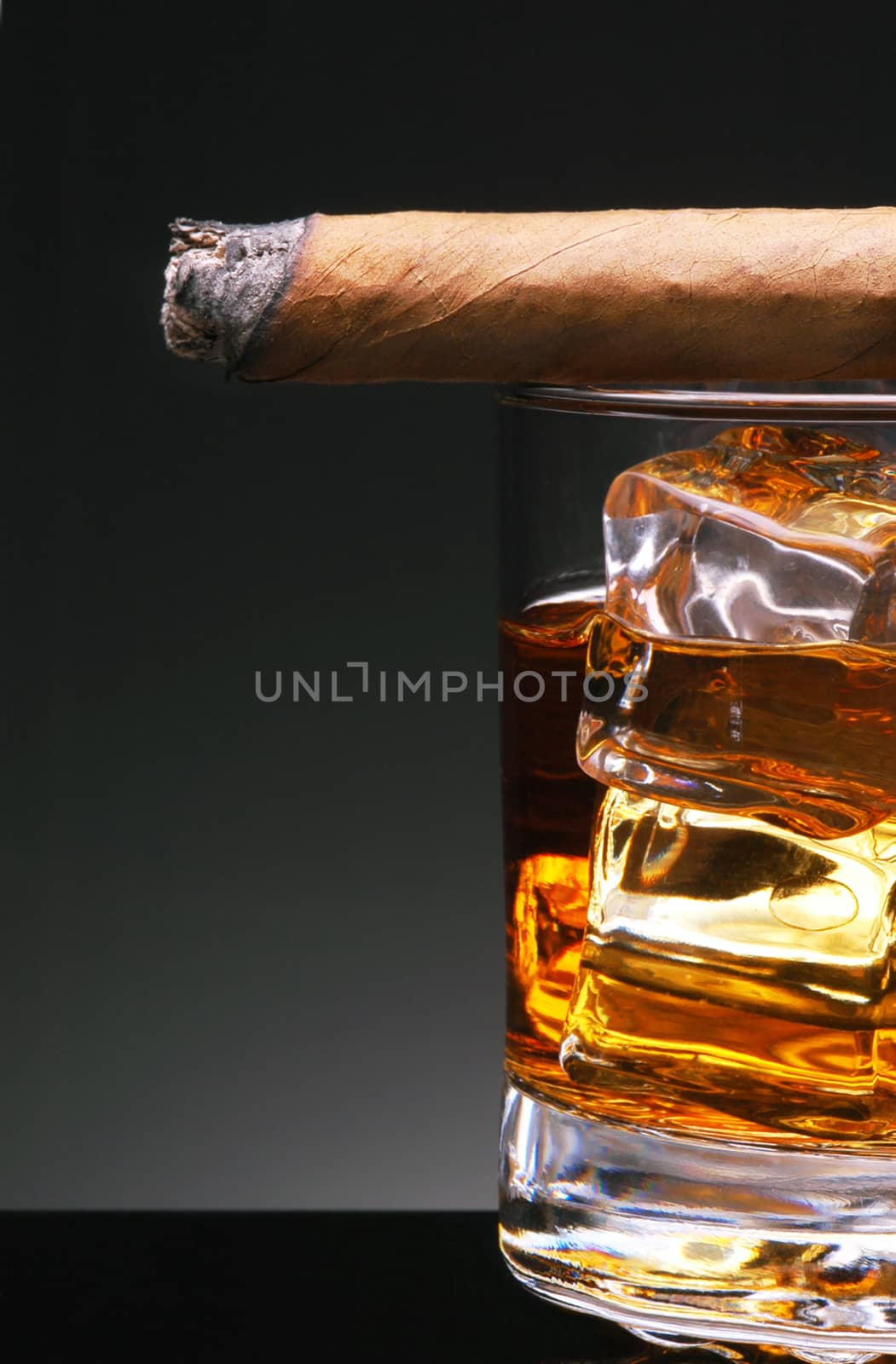 Closeup of  a lit cigar resting on top of a glass with ice cubes and whiskey, on a light to dark background.
