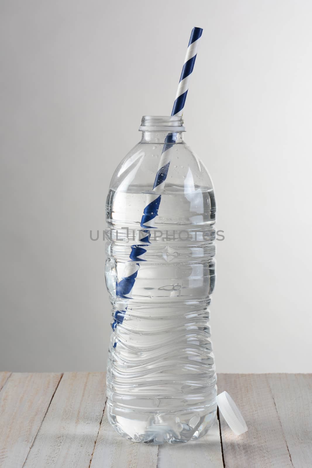 Closeup of a clear water bottle against a light to dark gray background. On a wood table the bottle has a blue and white striped drinking straw.