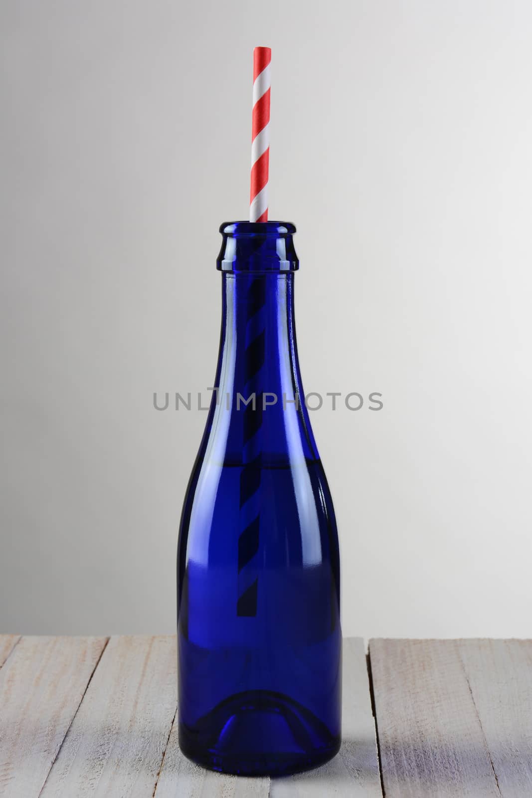 Closeup of a blue bottle with a red striped drinking straw. The bottle is on a rustic wood table with a light to dark gray background.