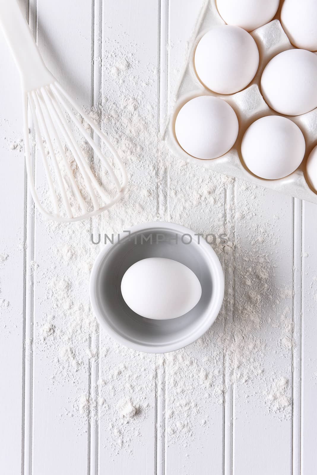 White on White baking still life. Top view of items for baking: eggs, flour and a whisk on a white beadboard surface. 