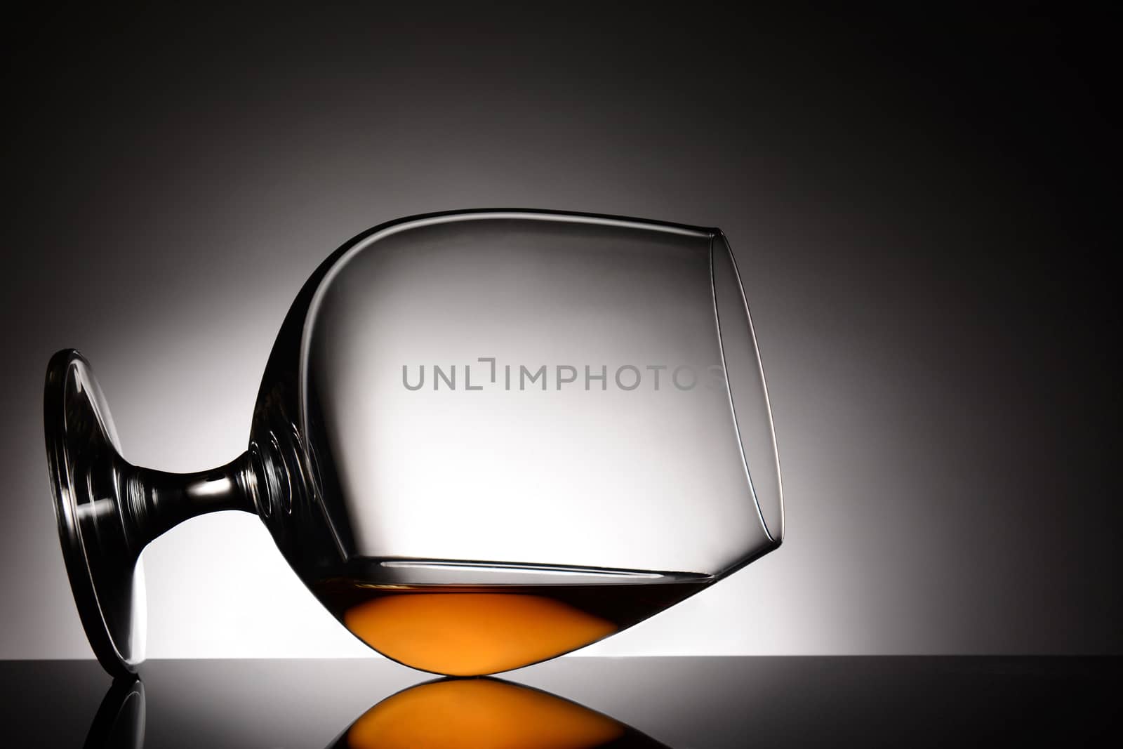 Closeup of a brandy snifter laying on it side. Horizontal format with a light to dark gray spot background. The glass is on a shiny black reflective surface.