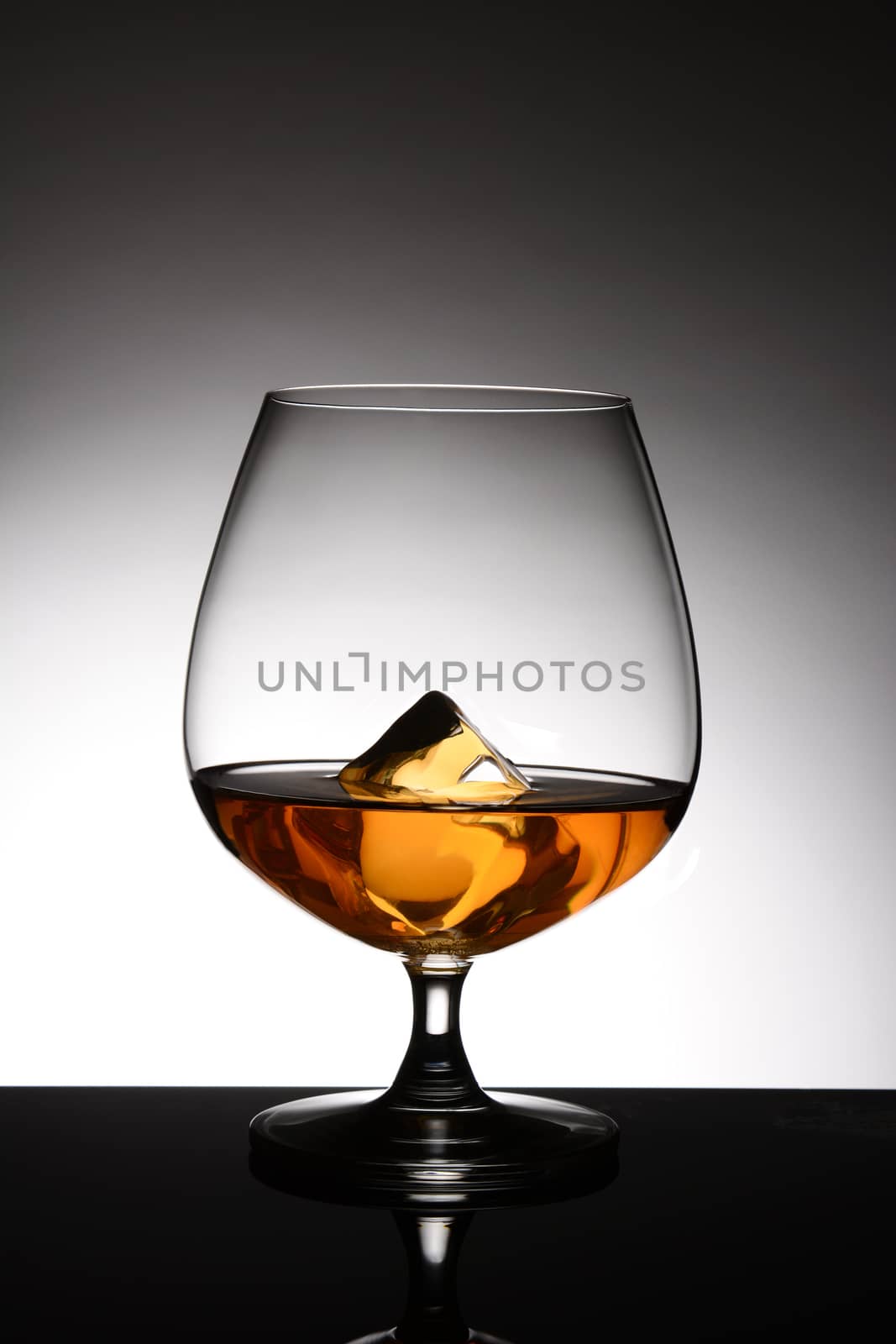 A brandy snifter with a single ice cube over a light to dark gray spot background. Vertical format.