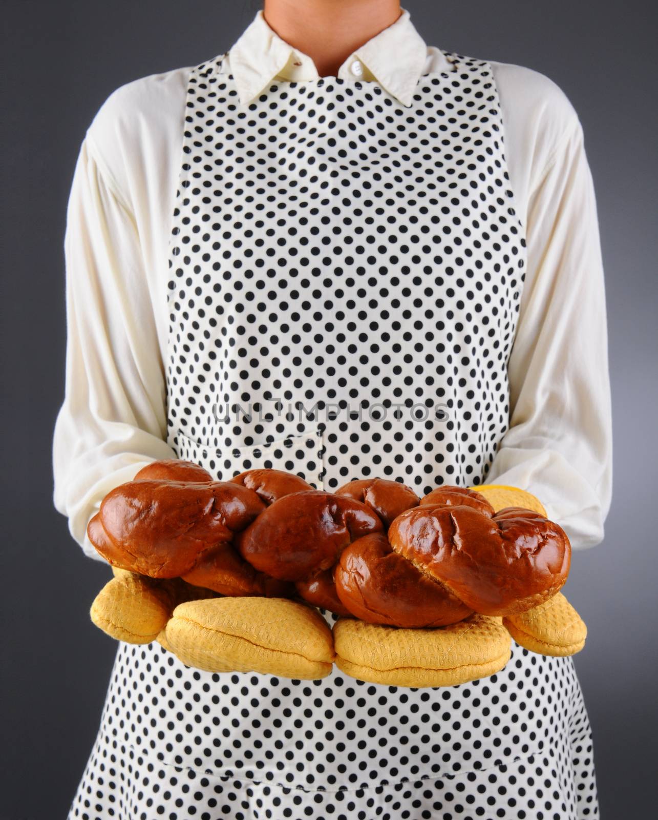 Closeup of a homemaker in an apron and oven mitts holding a freshly baked loaf of challah bread. Horizontal format over a light to dark background. Woman is unrecognizable. Shallow depth of field.