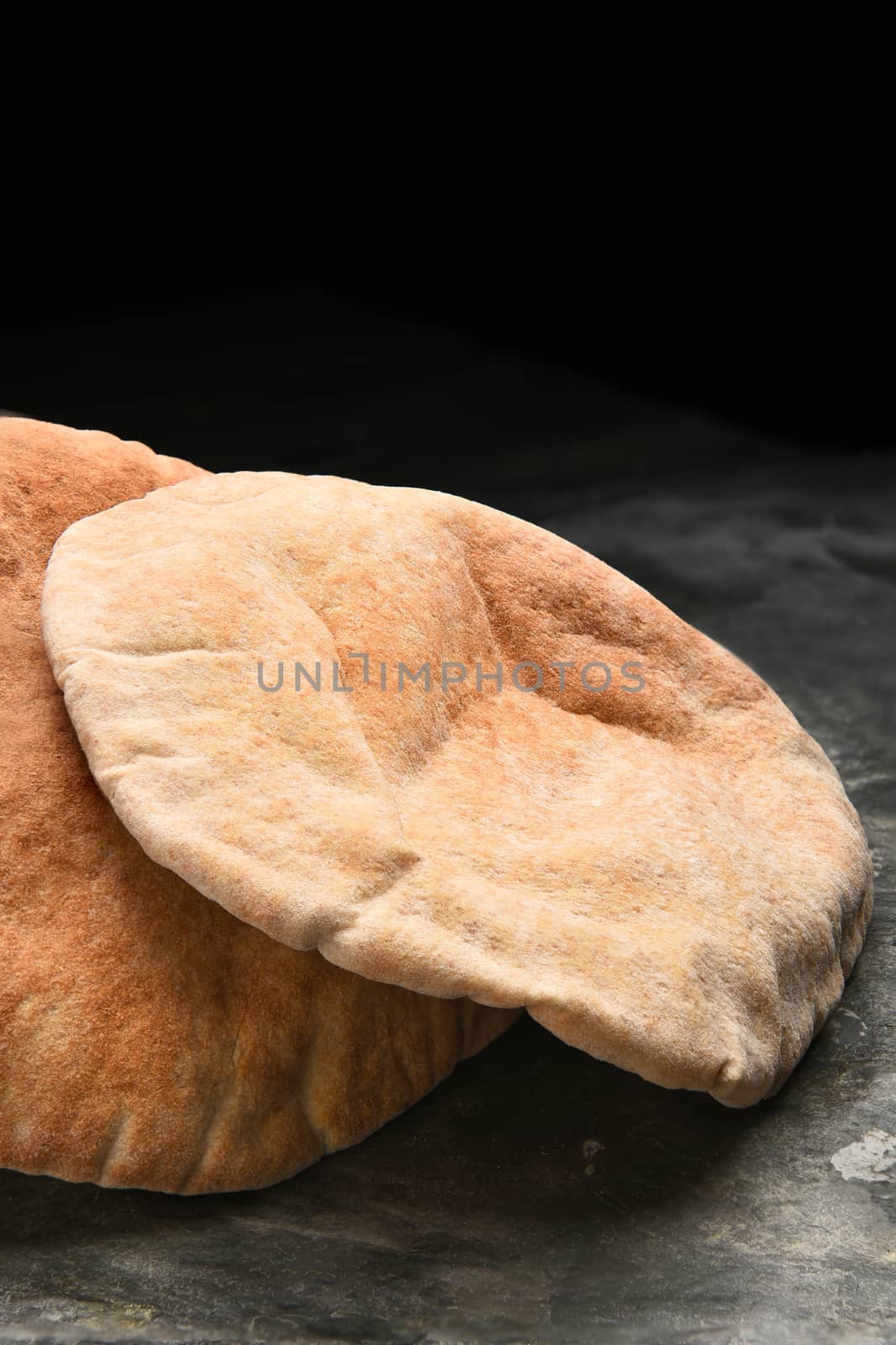 Vertical closeup of loaves of whole wheat pita bread on a slate surface and black background.