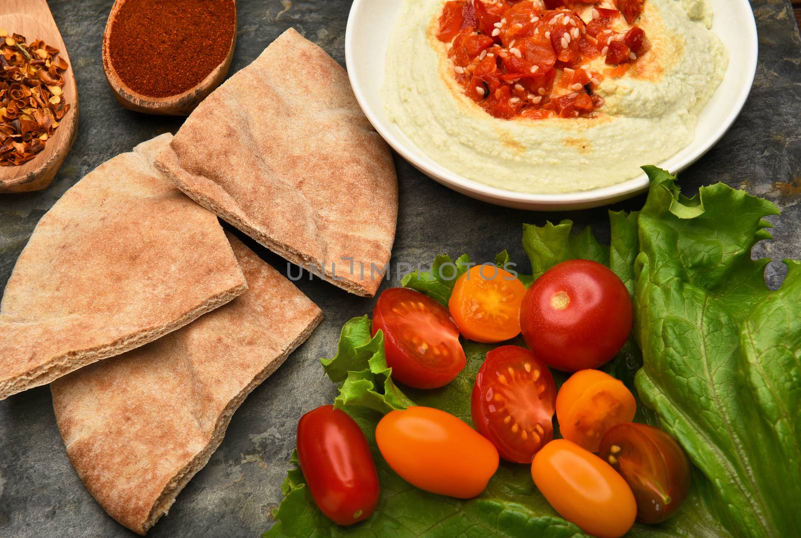 Top view of a bowl of edamame hummus and pita wedges on a slate surface. Lettuce leaf, tomatoes, and spices fill the scene.