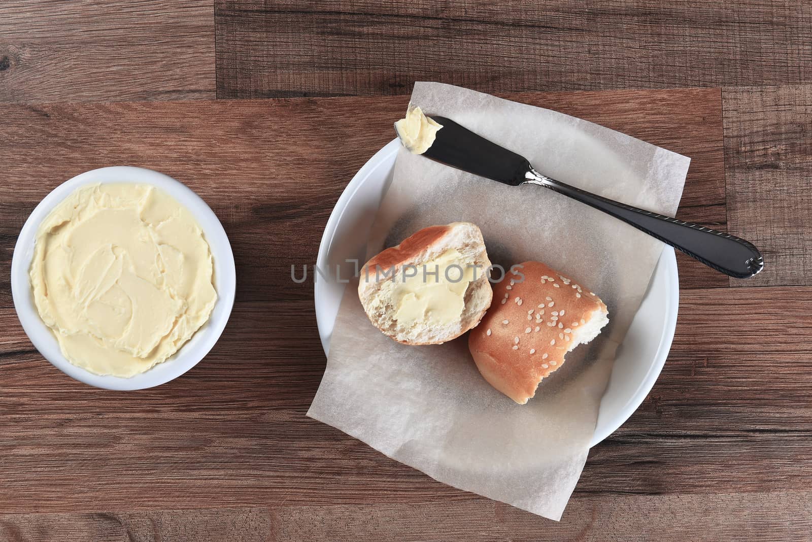 High angle view of a sesame dinner rolls broken in half and buttered. Next to the bread plate is a crock of butter with knife.