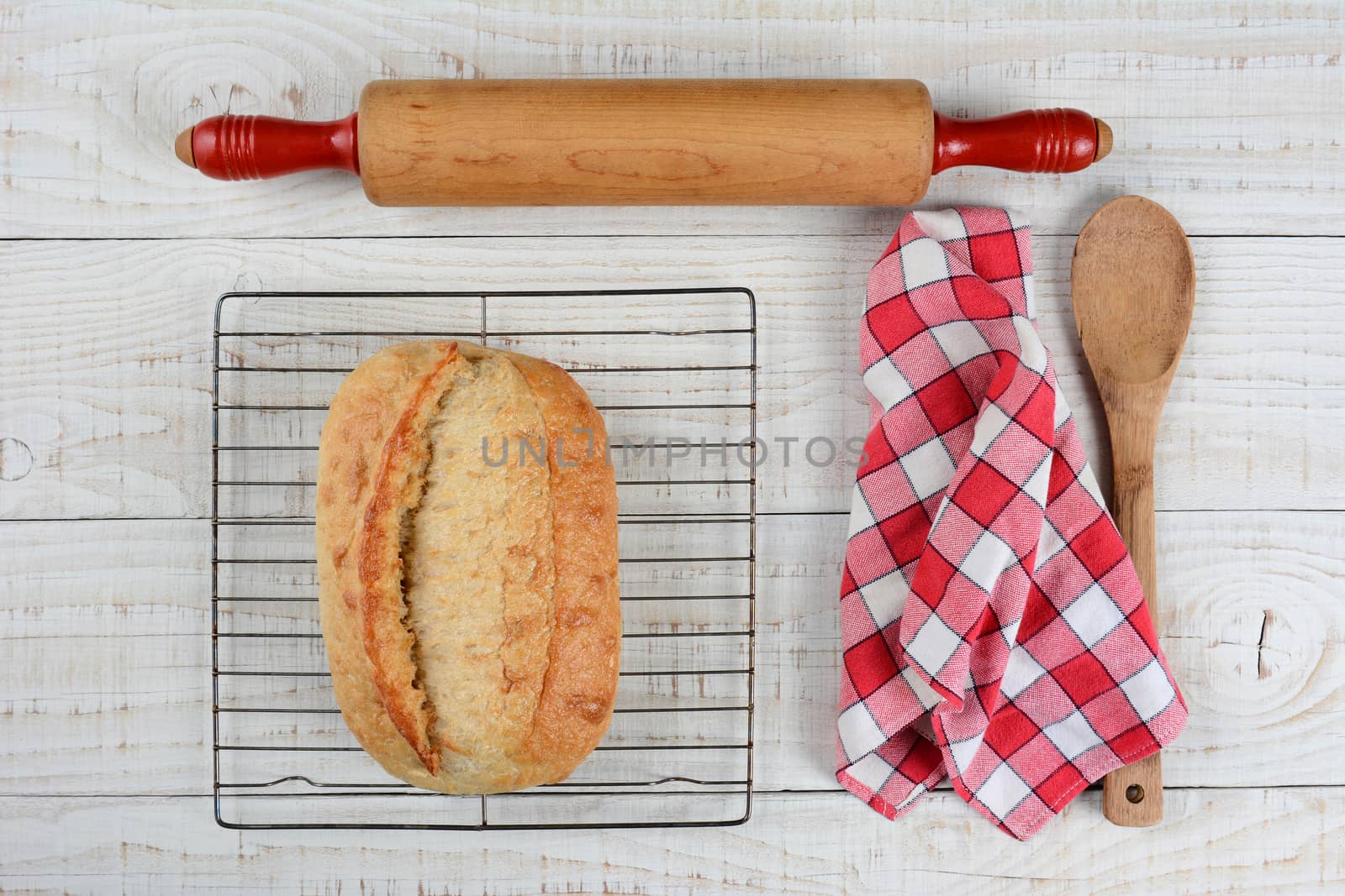 High angle shot of a fresh baked loaf of bread on a cooling rack with a rolling pin, red and white checkered napkin and wooden spoon, Horizontal format on a rustic white wood table.