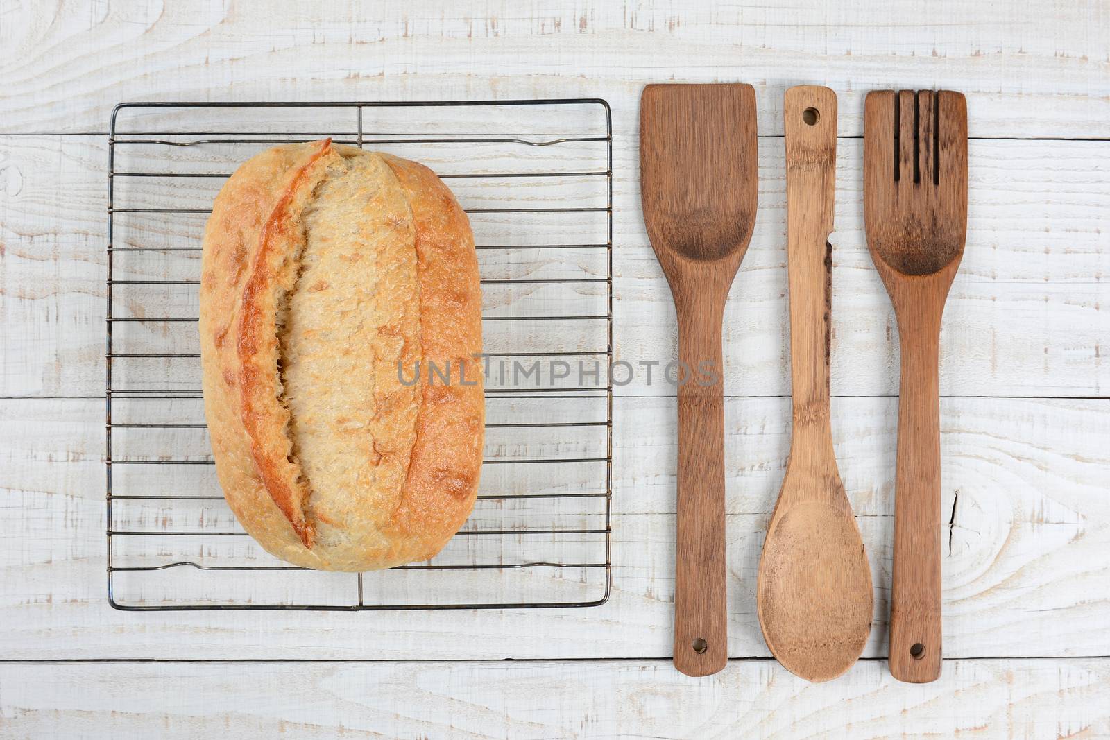 High angle shot of a fresh baked loaf of bread on a cooling rack with wooden spoon, spatual and fork. Horizontal format on a rustic white wood table.