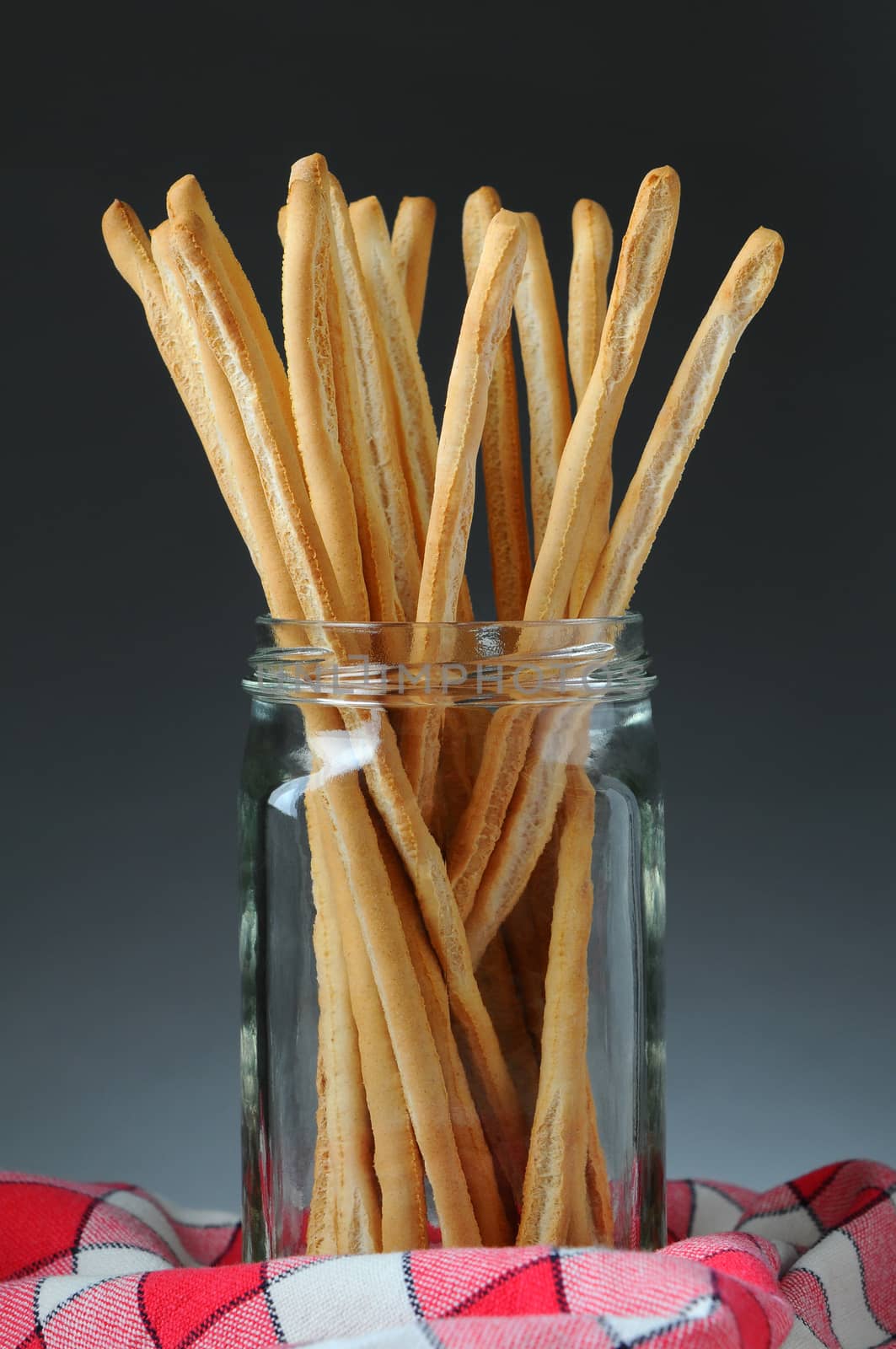 Closeup of a jar full of bread sticks on a checkered napkin with a light to dark gray background.